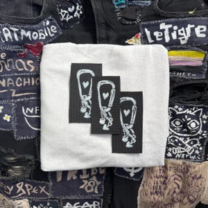 May include: Three black patches with white outlines of a climbing carabiner with a heart inside. The patches are sewn onto a white fabric background.