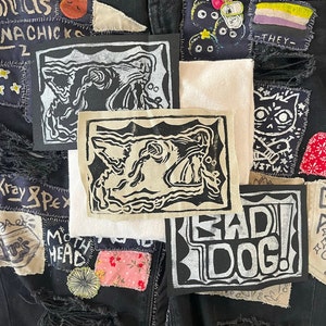 May include: A black denim jacket with various patches sewn on. The patches feature different designs, including a black and white dog head, a white cat head with a crossbones, and a white patch with the text "BAD! DOG!" in black.