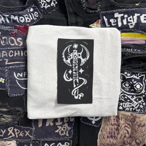 May include: A white fabric patch with a black background featuring a white stylized illustration of an axe with a curved blade and a handle with a knot at the top. The axe is surrounded by a white design that resembles a snake or a vine.