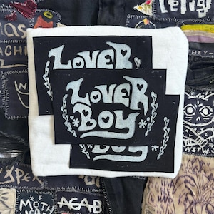 May include: Two black fabric patches with white lettering that reads "Lover Boy" on a white background. The patches are layered on top of each other.