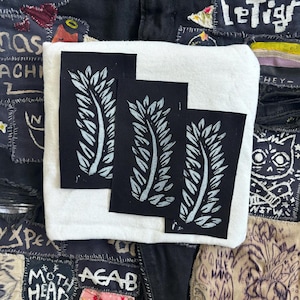 May include: Three black fabric patches with white leaf designs. The patches are sewn onto a white fabric background.