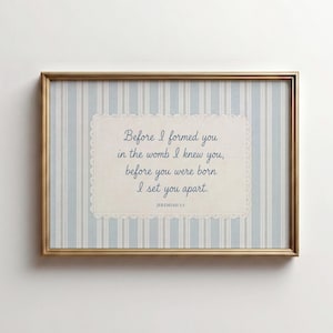 Puede incluir: Un cuadro rectangular con marco dorado y fondo de rayas verticales azul claro y blanco. En el centro, una tarjeta color crema con borde de encaje contiene el texto en azul: Before I formed you in the womb I knew you, before you were born I set you apart. JEREMIAH 1:5.