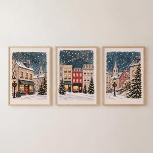 May include: A set of three framed wall art prints depicting winter town scenes at night. Each print features snow-covered buildings, evergreen trees, glowing street lamps, and string lights against a dark, starry, and snowy sky background.