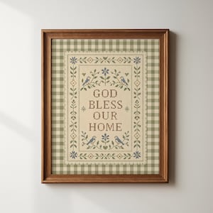 Op de afbeelding: Een ingelijste kruissteek met de tekst GOD BLESS OUR HOME in bruine letters. Het ontwerp bevat blauwe vogels, groene bladeren en kleine bloemen op een crèmekleurige achtergrond met een groen-witte vichy-ruitjesrand.