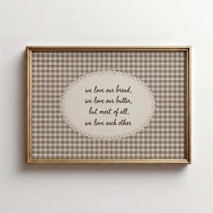 May include: A rectangular wall art piece in a gold frame featuring a beige and white gingham background. Centered is an oval cream-colored plaque with a scalloped edge containing the black text: we love our bread, we love our butter, but most of all, we love each other.