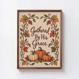 May include: A framed cross-stitch embroidery featuring the text Gathered By His Grace in dark brown script. The design is surrounded by a border of autumn-coloured maple leaves in shades of orange, yellow, and green, with three pumpkins at the base.