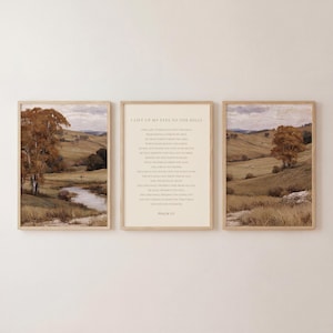 Peut inclure: Un ensemble de trois cadres muraux en bois clair. Les deux cadres extérieurs présentent une peinture de paysage avec des collines, des arbres aux tons dorés et un ruisseau. Le cadre central affiche le texte du Psaume 121 en police serif.