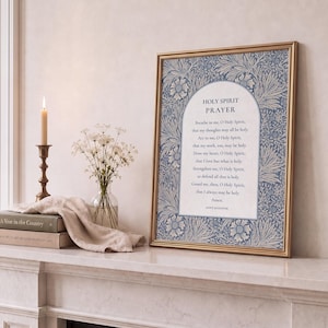 May include: A framed print with a gold border displays the "Holy Spirit Prayer" in a decorative font. The background features a blue and white floral pattern. A lit candle, flowers in a vase, and books are on the mantel.