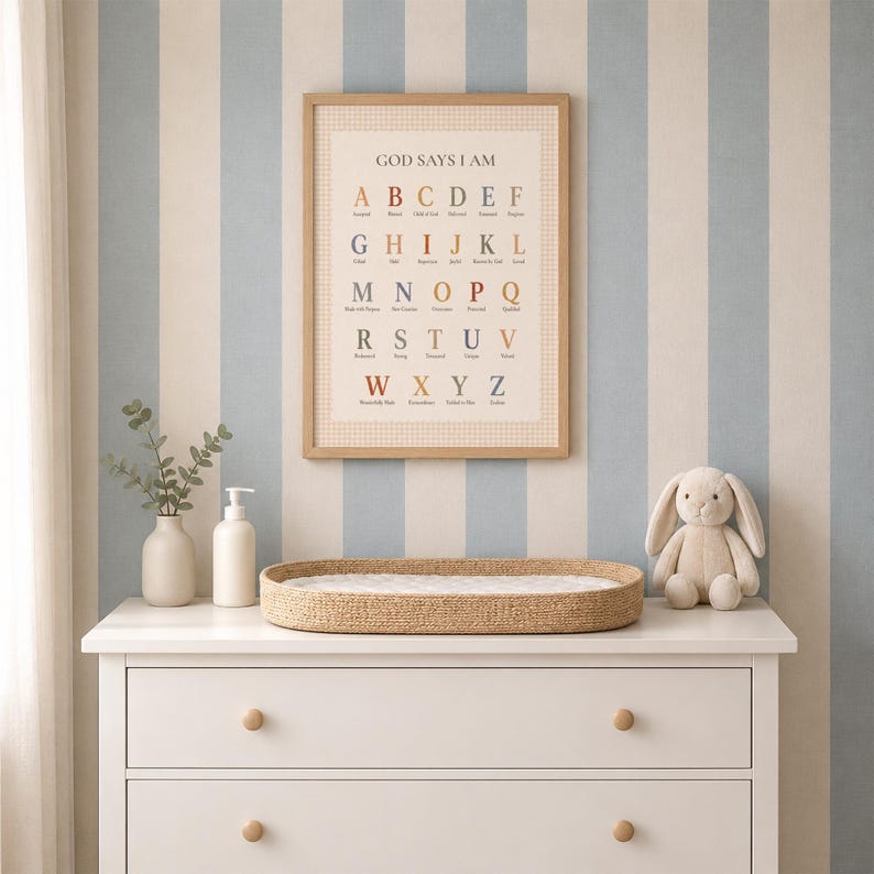 May include: A framed wall print titled GOD SAYS I AM featuring an alphabet chart with colorful letters and corresponding words, displayed above a white dresser with a woven changing pad, a white vase with greenery, and a plush rabbit toy.