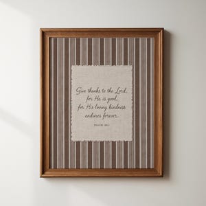 May include: A framed wall art piece featuring a rectangular piece of beige fabric with frayed edges centred on a brown and white striped background. The text reads: Give thanks to the Lord, for He is good, for His loving kindness endures forever. PSALM 136:1.