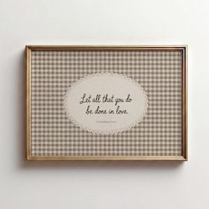 May include: A rectangular wall art piece in a gold frame featuring a beige and white gingham background. Centered is an oval cream-colored label with a scalloped edge containing the black text: Let all that you do be done in love. 1 Corinthians 16:14.