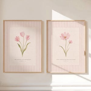 Pode incluir: Dois quadros decorativos com ilustrações florais em rosa claro sobre fundo creme com riscas verticais cor-de-rosa. A impressão da esquerda mostra três túlipas com o texto The Lord bless you and keep you, e a da direita dois cosmos com o texto I have called you by name; you are mine.