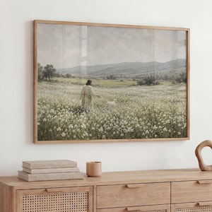 May include: A framed landscape painting depicting a figure in a long robe walking through a field of white flowers toward a small white lamb, with rolling hills and a muted sky in the background, presented in a light-toned wooden frame.