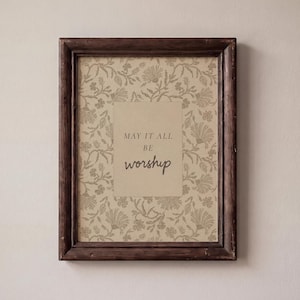 May include: A rectangular dark brown wooden frame containing a print with a beige background and a repeating floral pattern in a muted brown tone. Centered on the print is a rectangular beige block with the text MAY IT ALL BE worship.
