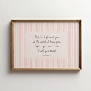 Può includere: Un quadro rettangolare con cornice dorata e sfondo a righe verticali rosa chiaro e bianco. Al centro, una targa rettangolare color crema con un bordo in pizzo smerlato riporta il testo: Before I formed you in the womb I knew you, before you were born I set you apart. JEREMIAH 1:5.