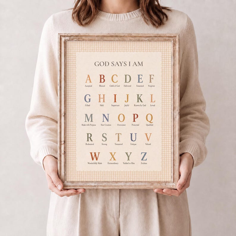 May include: A framed wall art print featuring the text GOD SAYS I AM at the top, followed by the alphabet from A to Z in a serif font. Each letter is paired with a corresponding word in a muted color palette, set against a light beige gingham background.