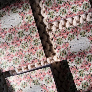 May include: Four notebooks with floral patterned covers in pink, green, and white. The covers feature a geometric design with the word "NOTEBOOK" in an oval frame. The notebooks are arranged on a textured, off-white surface.