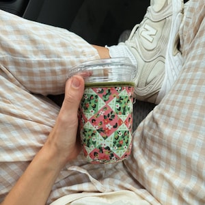 May include: A clear plastic cup with a green beverage is held in a floral patterned sleeve. The sleeve features a patchwork design with pink, green, and white floral elements. The person is wearing beige and white checkered pants and white sneakers.