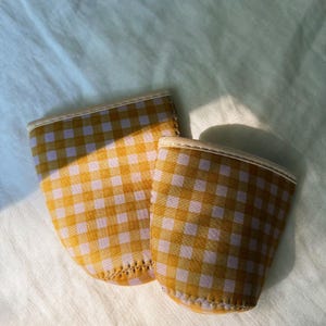 May include: Two yellow and white checkered cup sleeves with a cream-colored trim. The sleeves are made of a soft material and are designed to insulate beverages. The background is a light-colored fabric.