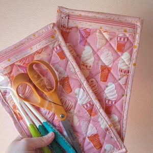 May include: Pink quilted zipper pouches with an ice cream cone pattern. One pouch holds orange scissors and crochet hooks. The other pouch is empty. The pouches are designed for craft supplies.
