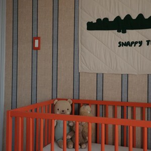 May include: An orange crib with a white mattress and two stuffed teddy bears. The crib is in front of a striped wall with a beige and blue pattern. A cream-colored quilt with a green alligator design and the words "Snappy Time" hangs on the wall.