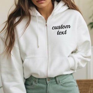 May include: A white zip-up hoodie with the words "custom text" in black script on the upper left chest. The hoodie has a drawstring hood, a full-length zipper, and two front pockets. The model is wearing light green jeans.