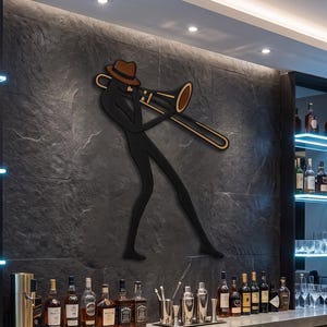 Op de afbeelding: Een zwart metalen wanddecoratie met een gestileerde figuur die een bruine hoed draagt en een goudkleurige trombone bespeelt. De figuur is bevestigd op een donkergrijze stenen muur boven een bar met diverse glazen flessen en baraccessoires.