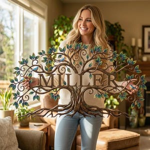 May include: A metal tree wall art piece with the name "MILLER" in the center. The tree has brown branches, green and blue leaves, and small blue bird accents. The art is held in front of a living room.