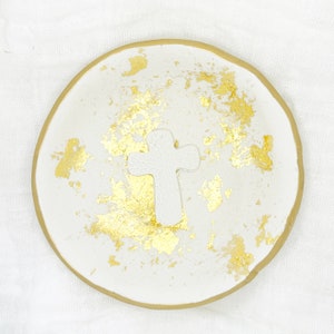 Puede incluir: Plato de cerámica blanco con borde dorado y motas doradas. Una cruz de cerámica blanca está en el centro del plato.