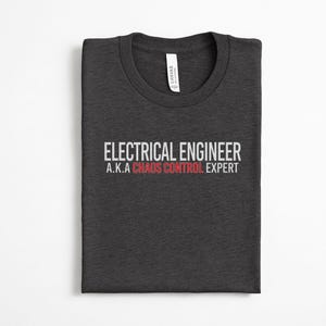 May include: A dark charcoal gray crew neck t-shirt featuring the white text ELECTRICAL ENGINEER and A.K.A. followed by the red text CHAOS CONTROL and the white text EXPERT printed on the front center of the garment with a visible brand label.