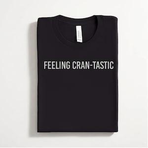 May include: A black crew neck t-shirt featuring the text FEELING CRAN-TASTIC printed in a light gray, distressed font across the chest. The garment includes a white interior neck label with the brand name Canvas clearly visible at the collar.