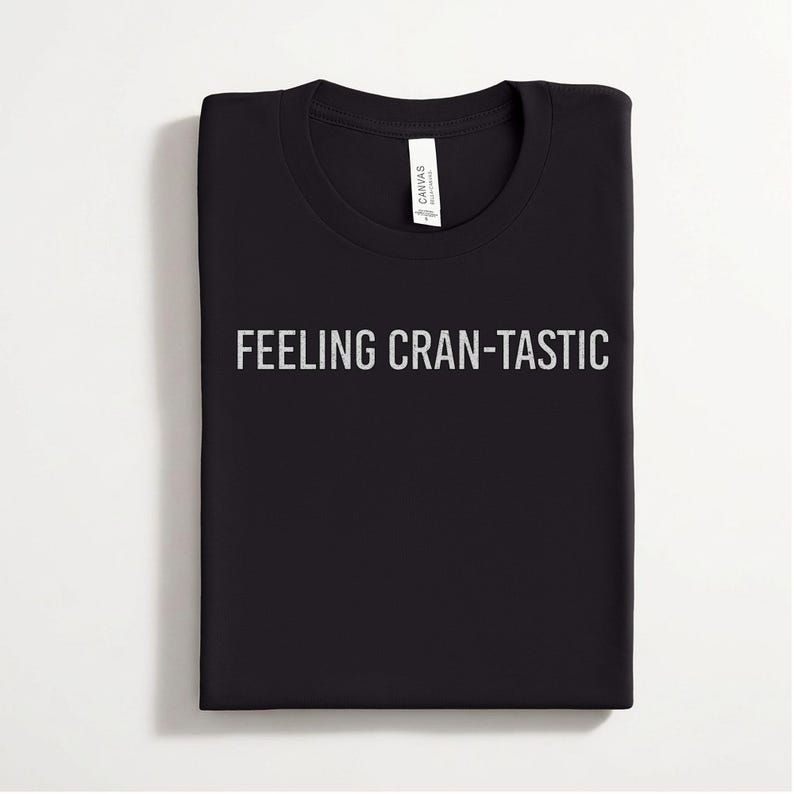 May include: A black crew neck t-shirt featuring the text FEELING CRAN-TASTIC printed in a light gray, distressed font across the chest. The garment includes a white interior neck label with the brand name Canvas clearly visible at the collar.