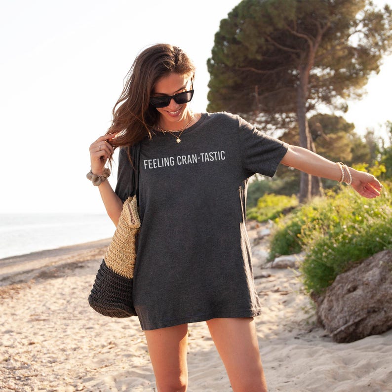 May include: A dark charcoal gray short-sleeve t-shirt featuring the white text "FEELING CRAN-TASTIC" printed across the chest. The shirt has a crew neckline and a relaxed fit, paired with a two-toned woven straw tote bag with a black base.