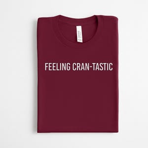 May include: A dark maroon crew neck t-shirt featuring the white text FEELING CRAN-TASTIC printed across the chest. The shirt has a standard short-sleeve design and includes a white brand label attached to the inside of the collar neckline.