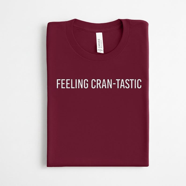 May include: A dark maroon crew neck t-shirt featuring the white text FEELING CRAN-TASTIC printed across the chest. The shirt has a standard short-sleeve design and includes a white brand label attached to the inside of the collar neckline.