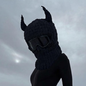 Puede incluir: Un pasamontañas negro tejido con cuernos adjuntos y gafas de esquí. La persona lleva un body negro de manga larga y pantalones negros. El fondo presenta montañas cubiertas de nieve y un cielo nublado.
