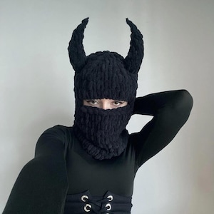 Puede incluir: Pasamontañas negro de punto con cuernos adjuntos. El pasamontañas cubre la cabeza y el cuello, con aberturas para los ojos y la boca. Los cuernos son curvos y texturizados. La persona lleva una camiseta negra de manga larga y un corsé.