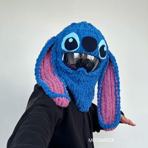 May include: A blue and pink crocheted hat resembling a cartoon character with large ears, eyes, and a mouth. The hat has a built-in visor and is worn over a black hoodie. The word "MOONWEEK" is visible on the right side.