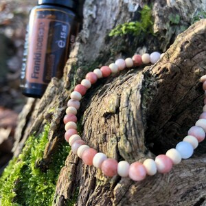 Könnte beinhalten: Ein rosa-weißes Perlenarmband mit einer kleinen Flasche Frankincense-ätherischem Öl. Das Armband besteht aus natürlichen Steinen und liegt auf einem mit Moos bewachsenen Baumstamm.