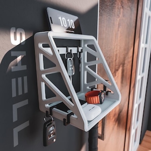 May include: A white wall-mounted organizer with a geometric design. It features a shelf, key hooks, and a slot for a tablet displaying "10 10". A watch with a red band, a pair of wireless earbuds, and keys are visible.