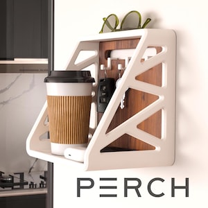 Puede incluir: Un organizador de pared blanco con un diseño geométrico, que contiene una taza de café, llaves y gafas de sol. El organizador tiene un panel trasero de madera y la palabra "PERCH" en la parte inferior.