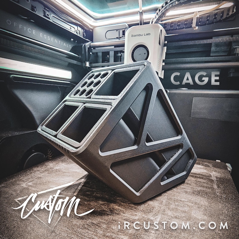 May include: A black 3D printed organiser with multiple compartments. The organiser is designed to hold office supplies and is labelled "OFFICE ESSENTIALS". The organiser is sitting on a wooden surface with the text "Custom" and "IRCUSTOM.COM" visible.