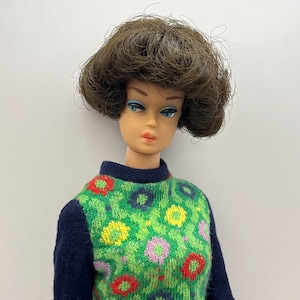 May include: A vintage doll with brown hair and blue eyes wearing a green and blue sweater with a floral pattern.