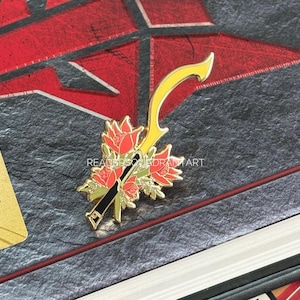 May include: A gold-colored pin featuring a yellow sword design with red and gold floral accents. The pin is set against a dark, textured background with red geometric shapes and a Barnes & Noble Exclusive card.