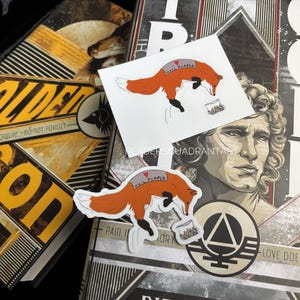 May include: Two stickers featuring a red fox with the text "Hail Alaper" on its back. The stickers are placed on a book with the title "The Golden Son" and the text "Hard and I do not forgive, I do not forget." The book also has a symbol of a triangle with a circle around it and the text "Pain is History" and "Love does not fade".