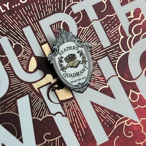 May include: A white enamel pin with a black and gold design. The pin features a shield with the words "Readers Quadrant" and "Read or Die" inside. The shield is surrounded by a floral design and the words "Readers Quadrant" are at the top.