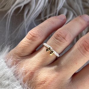 Könnte beinhalten: Ein goldfarbener Ring mit Schleife und einem weißen, perlenbesetzten Band. Der Ring wird an einem Finger getragen, im Hintergrund ein weißer, flauschiger Ärmel. Der Ring ist ein zartes Schmuckstück.