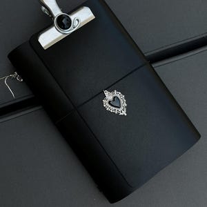 May include: A black leather journal cover featuring a silver metal clip at the top with a black circular inlay. A thin black elastic band wraps around the centre, holding a silver-toned heart-shaped charm with a black stone centre.
