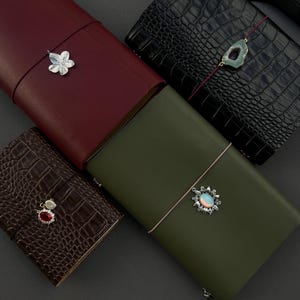 May include: Four rectangular journals with elastic closures and decorative charms. One is smooth burgundy, one is smooth olive green, one is black with a crocodile texture, and one is dark brown with a crocodile texture and a red stone charm.