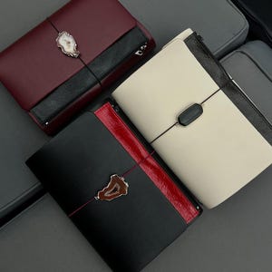 Peut inclure: Trois carnets rectangulaires avec fermetures élastiques. L'un est bordeaux foncé avec un accent noir et une pierre d'agate blanche. L'un est crème avec un accent noir et une pierre noire. L'un est noir avec un accent rouge et une agate brune.